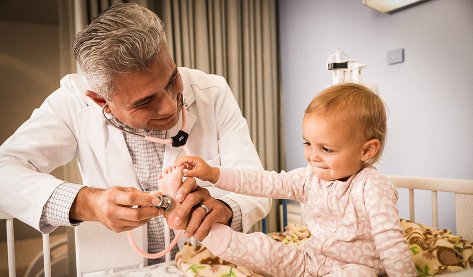Older male physician looks at a child patient who is playing with his stethoscope
